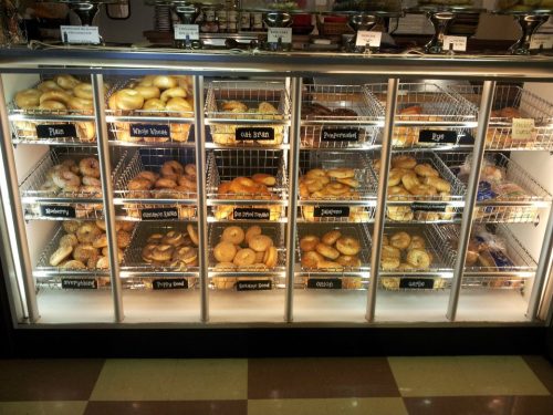 A display case filled with various types of bagels, labeled with flavors like plain, blueberry, and everything.