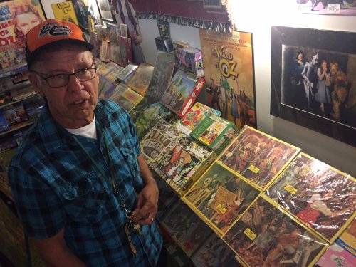 An older man in a cap stands beside a display of vintage magazines and memorabilia in a colorful room.