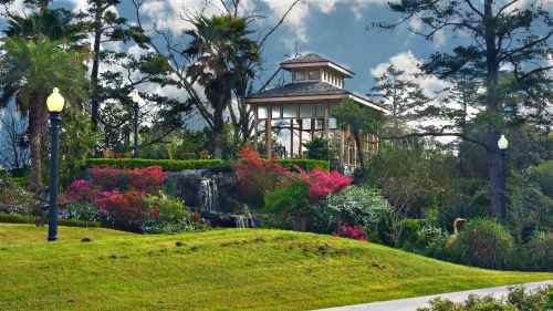 A scenic garden with a gazebo, colorful flowers, and palm trees under a cloudy sky.