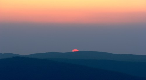 A serene sunset over rolling hills, with the sun partially hidden on the horizon, casting warm hues in the sky.