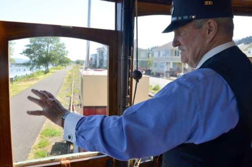 A conductor waves from inside a vintage streetcar, with a scenic path and houses visible outside the window.