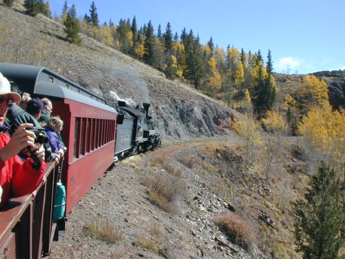 A vintage steam train travels along a scenic mountain route, surrounded by autumn foliage and rocky terrain.
