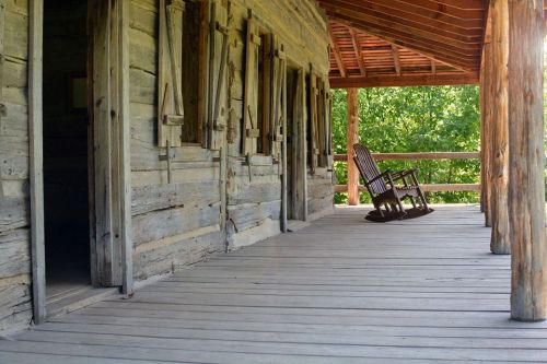 A rustic wooden porch with a rocking chair, surrounded by greenery and sunlight filtering through the trees.