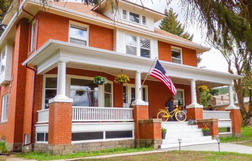 A red brick house with a porch, American flag, and a yellow bicycle parked outside on a sunny day.