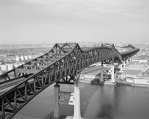 A black and white image of a large, arched bridge spanning over a body of water, with a city skyline in the background.