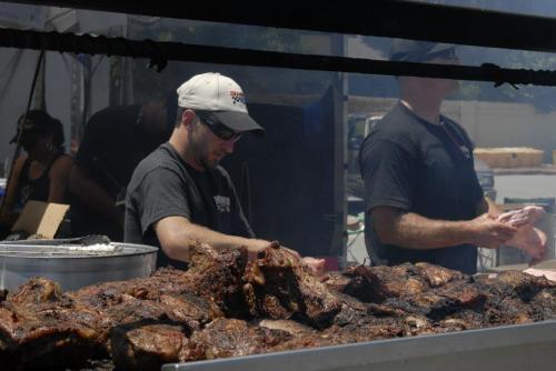 Two men grilling meat at a barbecue event, with smoke rising and a variety of meats on the grill.
