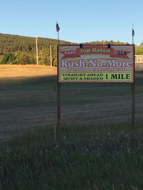 Sign for "Rush-No-More RV Park & Campground," indicating it's 1 mile ahead, with a scenic background of hills.