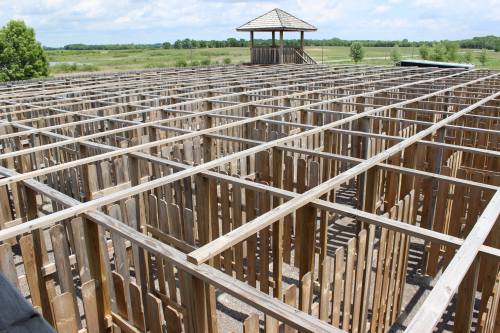 A wooden maze structure viewed from above, with a lookout tower in the background and green fields beyond.