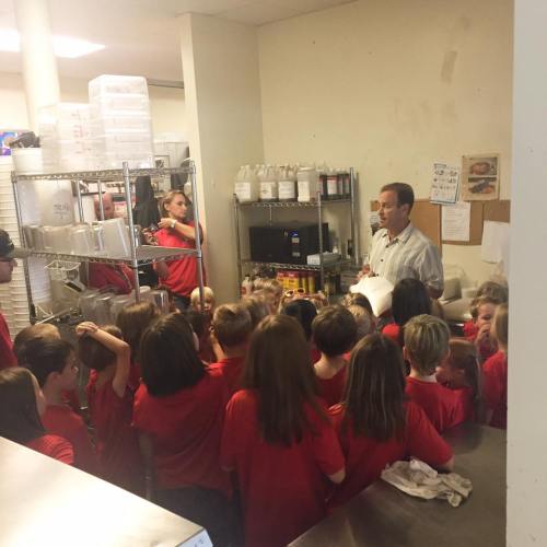 A group of children in red shirts gathers around an adult in a kitchen, listening attentively to a demonstration.