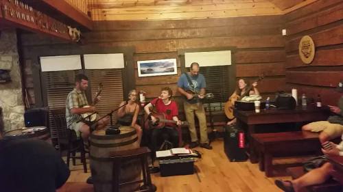 A group of musicians performs in a cozy wooden room, playing guitars and banjos, with an audience enjoying the music.