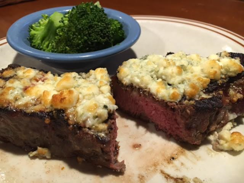 A plate with two slices of steak topped with crumbled cheese, served with a side of broccoli in a blue bowl.