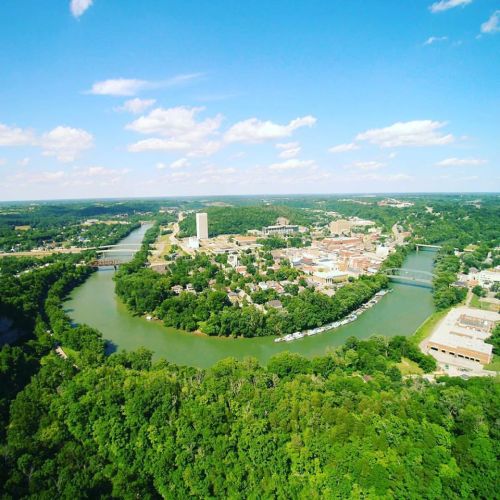 Aerial view of a lush green landscape with a winding river and a small town under a bright blue sky with clouds.