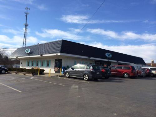 A one-story building with a black roof and blue windows, surrounded by parked cars under a clear sky.