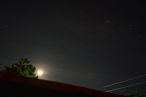 A night sky filled with stars, featuring a bright moon partially obscured by clouds, above a silhouetted rooftop and tree.