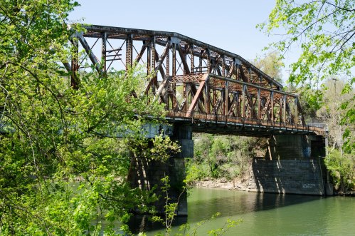A rusty metal bridge spans a calm river, surrounded by lush green trees and a clear blue sky.