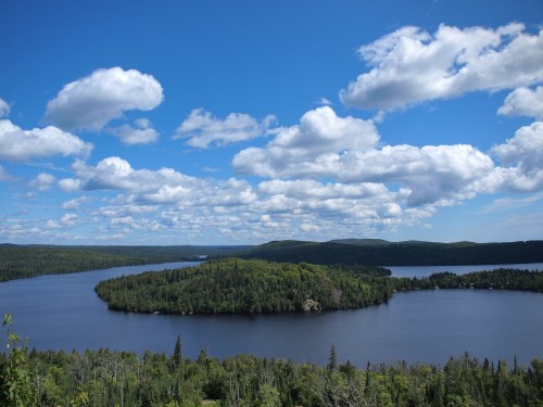 A serene lake surrounded by lush green forests under a bright blue sky with fluffy white clouds.