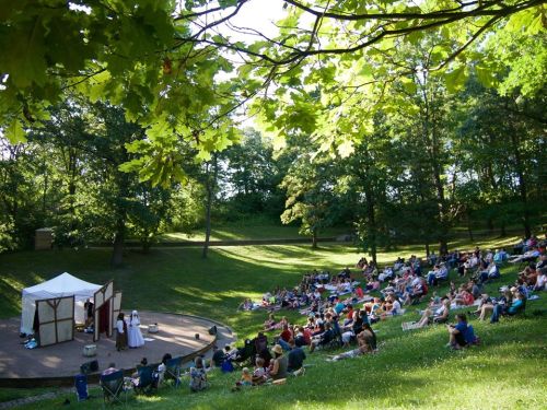 A crowd enjoys an outdoor performance in a lush green park, seated on blankets and chairs under a sunny sky.