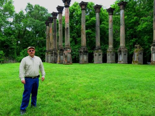 A man stands in front of tall, weathered columns in a green field, surrounded by trees.