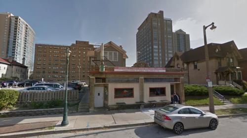 A pizza restaurant with a red awning, surrounded by tall buildings and greenery on a city street.