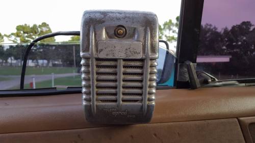 A vintage metal microphone mounted on a dashboard, with a blurred outdoor background.