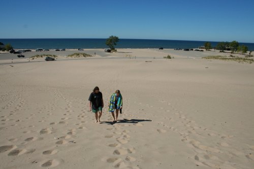 Two people walk barefoot on a sandy beach, with a clear blue sky and ocean in the background. Cars are parked nearby.