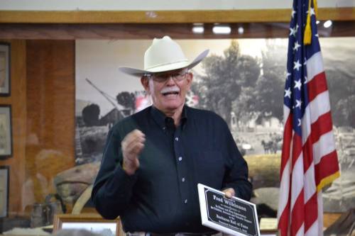 A man in a white cowboy hat speaks at an event, holding a plaque, with a U.S. flag and historical photos in the background.