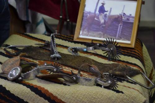 A collection of vintage spurs on a woven blanket, with a framed photo of a cowboy on horseback in the background.