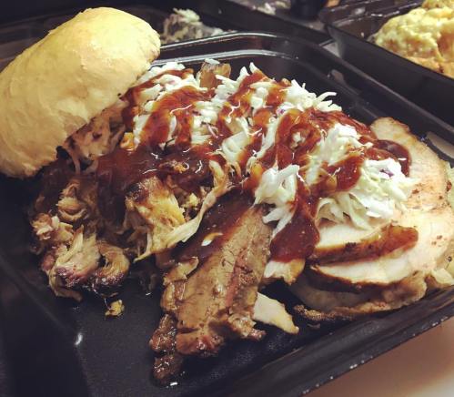 A hearty barbecue sandwich with pulled pork, sliced meat, coleslaw, and barbecue sauce on a soft bun, served in a black container.