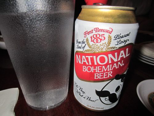 A can of National Bohemian Beer next to a frosted glass of water on a table.