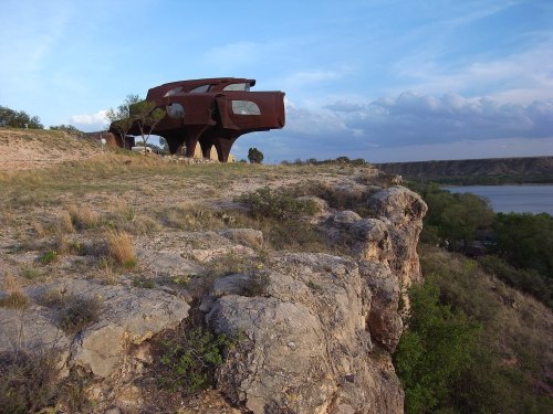 A unique, modern building perched on a rocky cliff overlooking a river and lush greenery under a blue sky.