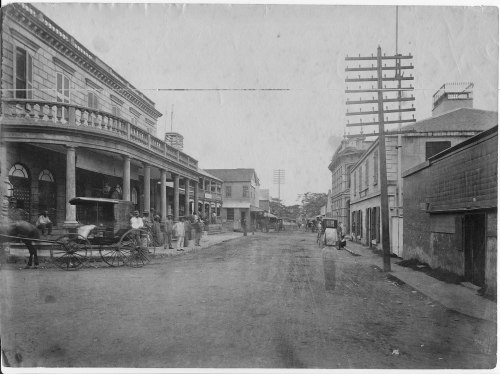 A historic street scene featuring horse-drawn carriages, buildings, and early telephone poles.