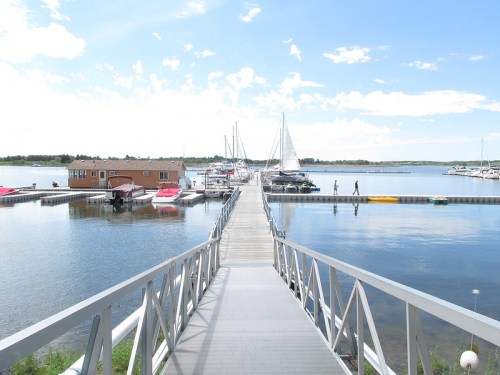 A wooden dock extends over calm water, lined with boats and a small building, under a bright blue sky.