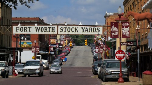 A view of Fort Worth Stockyards with a large sign, vintage cars, and historic buildings under a blue sky.