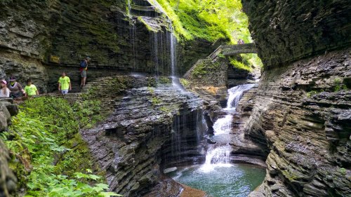 A scenic gorge with waterfalls, lush greenery, and visitors exploring the rocky terrain along a winding path.