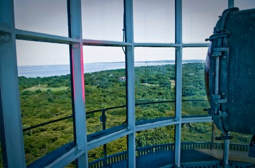 View from inside a lighthouse, showcasing lush greenery and a distant coastline through large glass panes.