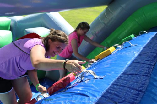 Two women in pink shirts climb an inflatable structure, reaching for colorful handles. Bright, sunny day outdoors.
