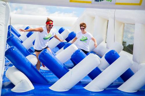 Two women in white shirts navigate an inflatable obstacle course with blue and white structures, smiling and having fun.