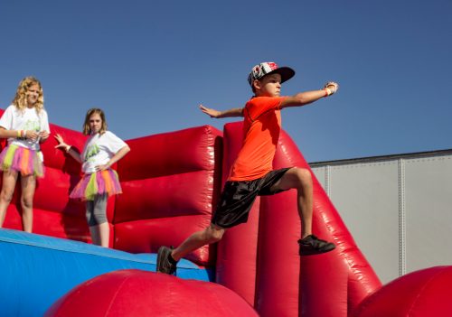 A boy in an orange shirt jumps on a colorful inflatable structure while two girls watch from behind.