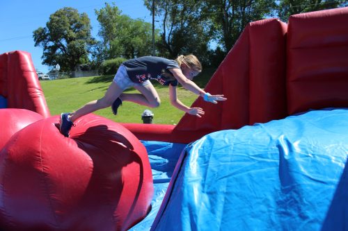 A child jumps across a large inflatable obstacle in a sunny outdoor setting.