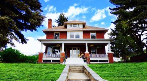 A large brick house with a front porch, surrounded by greenery and trees, under a blue sky with clouds.