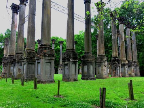 Ruins of tall, weathered columns surrounded by lush green grass and trees, creating a serene, historical atmosphere.