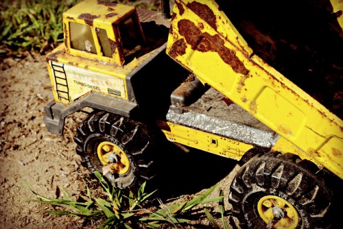 A rusty yellow toy dump truck sits on dirt, surrounded by green grass, showing signs of wear and age.
