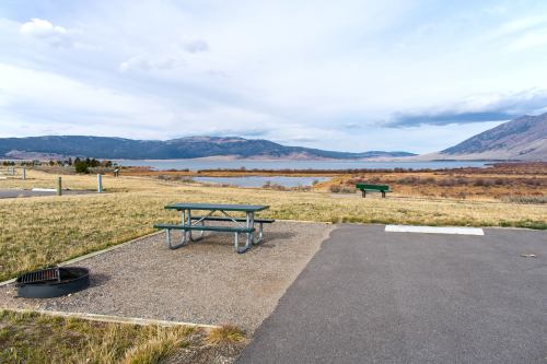 A picnic area with tables near a lake, surrounded by mountains and grassy fields under a cloudy sky.