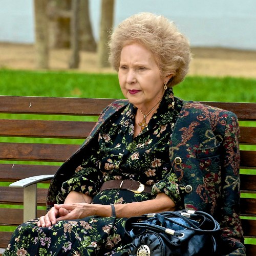 An elderly woman with curly hair sits on a bench, wearing a floral dress and a patterned jacket, holding a handbag.