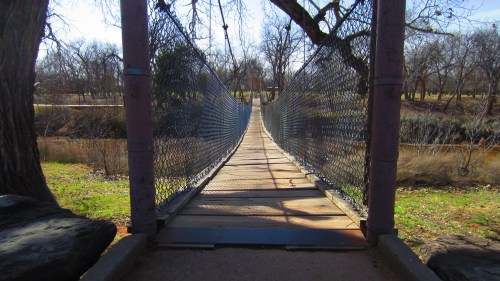 A wooden suspension bridge stretches over a river, surrounded by trees and grass in a serene park setting.