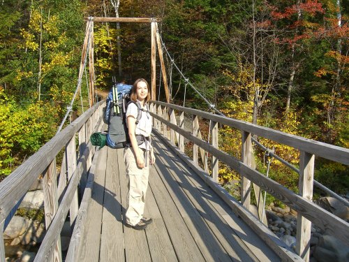 A hiker with a backpack stands on a wooden bridge surrounded by colorful autumn trees.