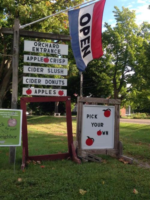A sign at an orchard entrance with "Open" flag, listing apple products and "Pick Your Own" options.