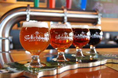 Four glasses of craft beer in a row, featuring different colors and styles, on a wooden tasting paddle.