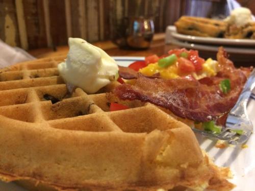 A close-up of a waffle topped with butter, alongside a plate of scrambled eggs, bacon, and diced vegetables.