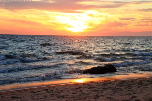 A vibrant sunset over the ocean, with waves gently lapping at the shore and a rock partially submerged in the sand.